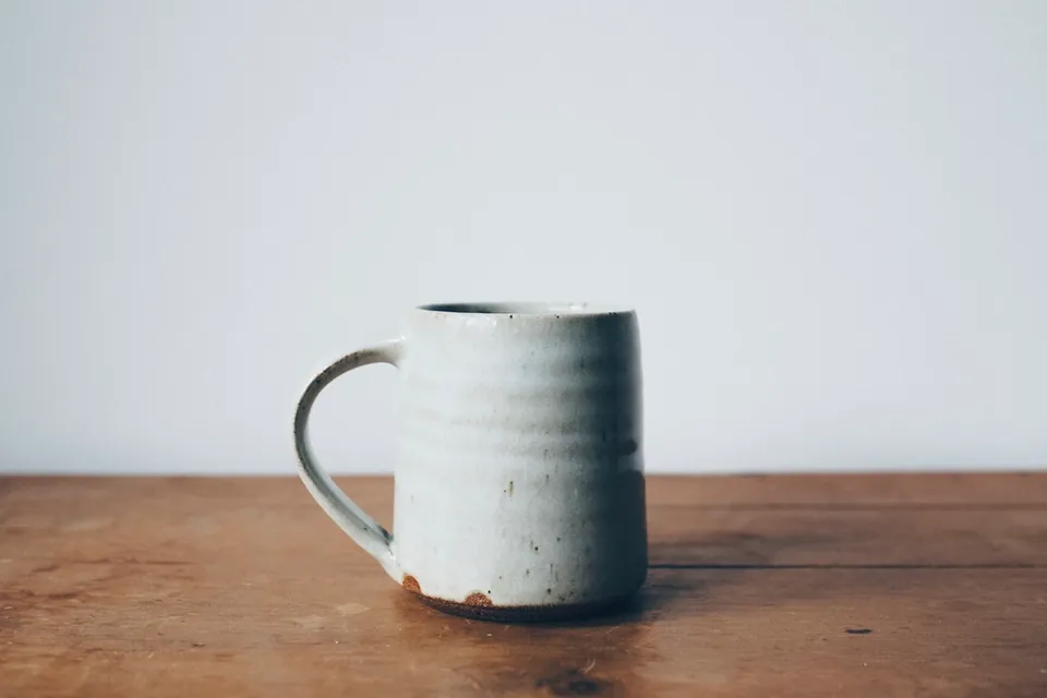 A handmade pale ceramic mug standing on a wooden table against a white wall