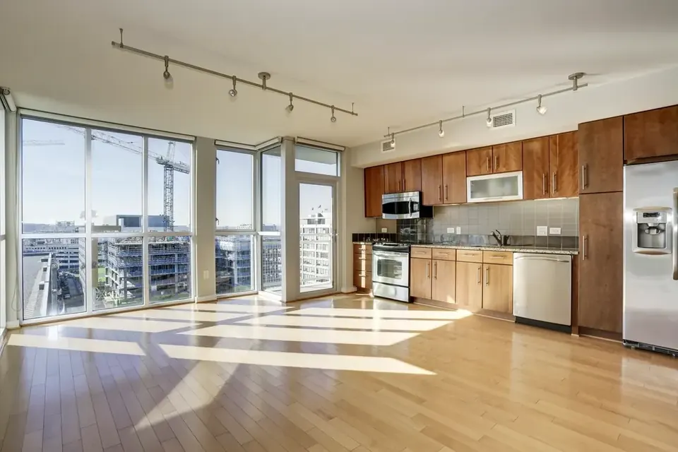 An empty flat with floor-to-ceiling windows, sunlight on a wooden floor and a fitted kitchen along one wall