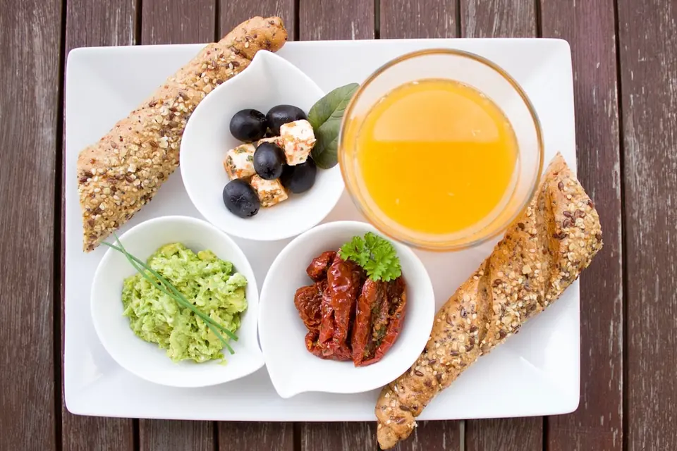 A breakfast plate of seeded bread, orange juice, olives, guacamole and sun-dried tomatoes